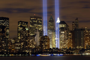 Photograph of a nighttime city skyline featuring two vertical beams of light representing the Tribute in Light memorial in New York City. The scene highlights illuminated skyscrapers with reflections on water in the foreground, symbolizing remembrance of the September 11 attacks.