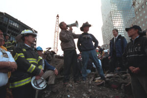Photograph showing emergency responders and officials gathered at a disaster site with debris and rubble, with one man using a megaphone to address the group. Background includes construction crane and tall buildings, highlighting coordination efforts during a rescue or recovery operation.