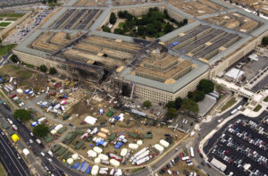 Aerial photograph of the Pentagon showing extensive damage to one side caused by a large impact. Surrounding area contains numerous emergency response tents, vehicles, and personnel indicating an ongoing rescue and recovery operation.