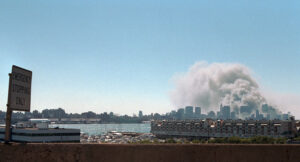 Photograph showing a city skyline with a large plume of smoke rising from multiple buildings, indicating a major fire or explosion. Foreground includes a waterfront marina and a sign reading "Emergency Stopping Only."
