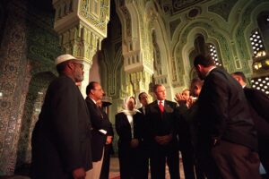 Photograph showing a group of people, including a man in a suit and a woman wearing a headscarf, standing inside an ornately decorated mosque with intricate green and gold patterns on arches and walls. The setting highlights cultural and religious architecture, with individuals engaged in conversation, suggesting a formal or diplomatic visit.